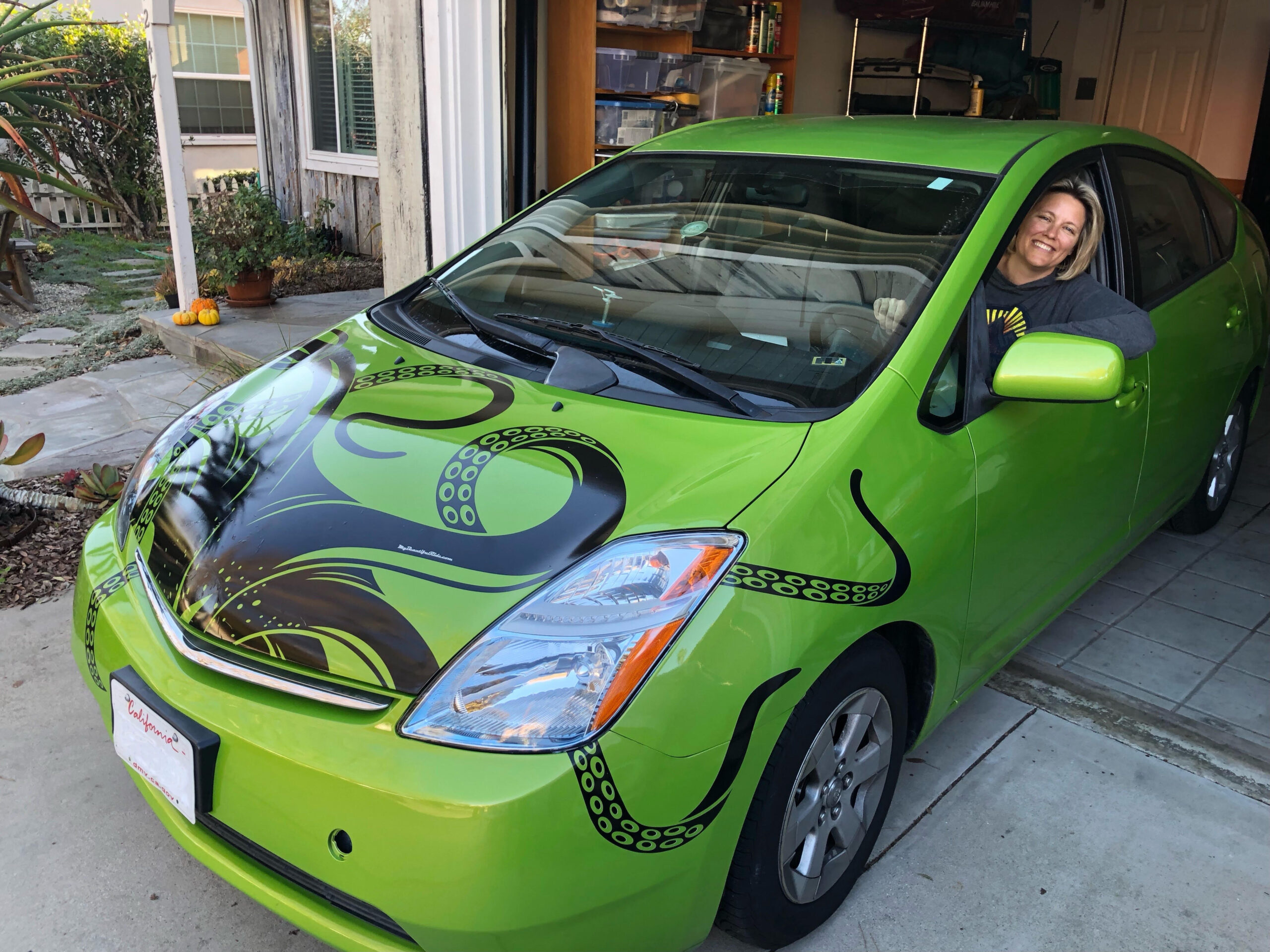 Blonde woman sitting in the driver's seat of a bright green prius with a giant kraken or octopus climing out of the hood engine compartment with tentacles down the sides of the car